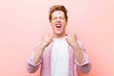 young red head man shouting triumphantly, laughing and feeling happy and excited while celebrating success against pink wall