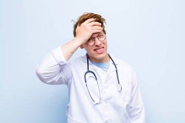 young red head doctor panicking over a forgotten deadline, feeling stressed, having to cover up a mess or mistake against blue wall