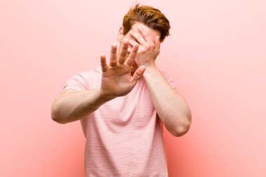 Genç kızıl saçlı adam elini yüzünü örtüyor ve diğer elini kamerayı durdurmak için öne koyuyor. Pembe duvara karşı fotoğraf ya da resim çekmeyi reddediyor.