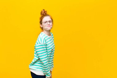 young red head woman with a goofy, crazy, surprised expression, puffing cheeks, feeling stuffed, fat and full of food against orange wall