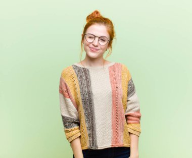 Genç ve güzel kızıl saçlı kadın mutlu ve arkadaş canlısı görünüyor, yeşil duvara karşı pozitif bir tavırla sana gülümsüyor ve göz kırpıyor.