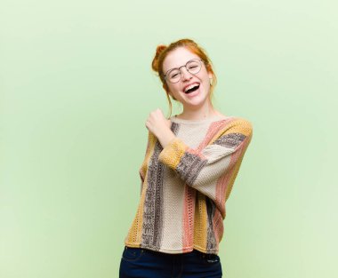 Genç, güzel kızıl saçlı kadın mutlu, pozitif ve başarılı hissediyor, meydan okumayla karşılaştığında motive oluyor veya yeşil duvara karşı iyi sonuçları kutluyor.