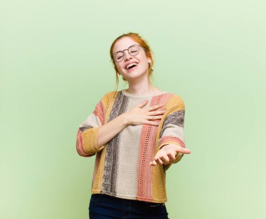 Genç ve güzel kızıl saçlı kadın mutlu ve aşık, bir eli kalbinin yanında gülümsüyor ve diğeri yeşil duvara yaslanmış.