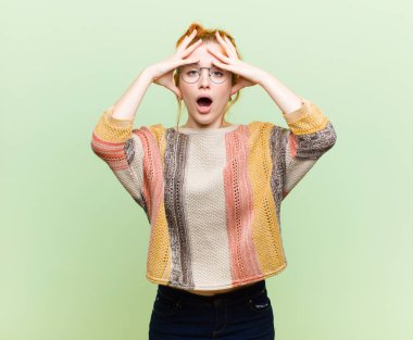 young pretty red head woman feeling horrified and shocked, raising hands to head and panicking at a mistake against green wall