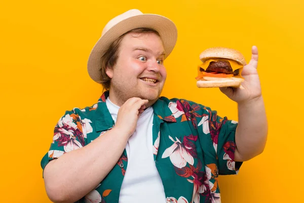 young big size man with a cheese burger against flat wall