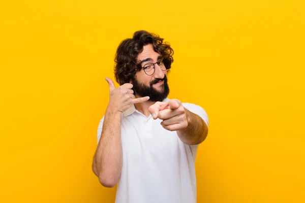 young crazy man smiling cheerfully and pointing to camera while making a call you later gesture, talking on phone against yellow wall