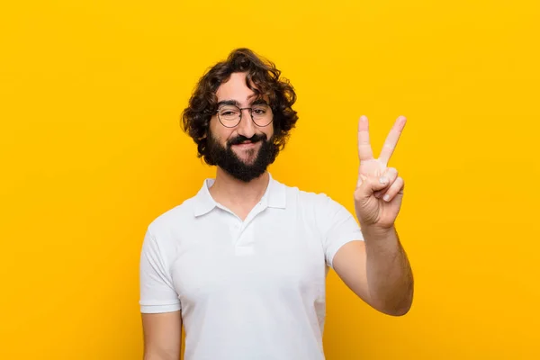 young crazy man smiling and looking friendly, showing number two or second with hand forward, counting down against yellow wall