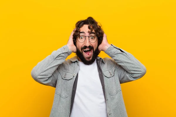 young crazy man raising hands to head, open-mouthed, feeling extremely lucky, surprised, excited and happy against yellow wall