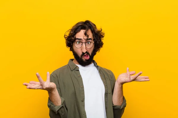 young crazy man shrugging with a dumb, crazy, confused, puzzled expression, feeling annoyed and clueless against yellow wall