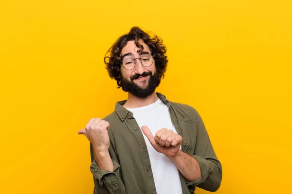 young crazy man smiling cheerfully and casually pointing to copy space on the side, feeling happy and satisfied against yellow wall