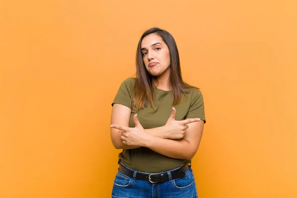 young pretty woman looking puzzled and confused, insecure and pointing in opposite directions with doubts isolated against orange wall