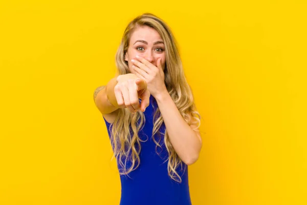 young blonde woman laughing at you, pointing to camera and making fun of or mocking you against yellow wall