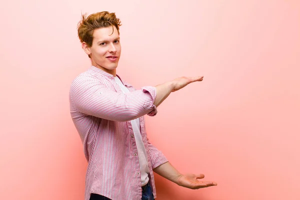 young red head man holding an object with both hands on side copy space, showing, offering or advertising an object against pink wall