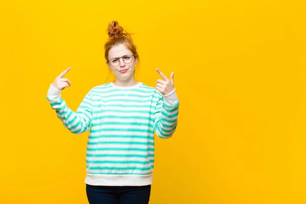 young red head woman with a bad attitude looking proud and aggressive, pointing upwards or making fun sign with hands against orange wall