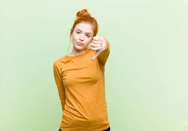 young pretty red head woman feeling cross, angry, annoyed, disappointed or displeased, showing thumbs down with a serious look against green wall