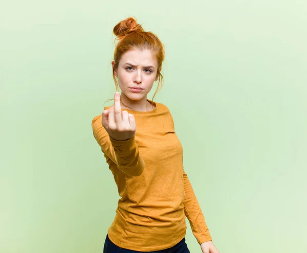 young pretty red head woman feeling angry, annoyed, rebellious and aggressive, flipping the middle finger, fighting back against green wall