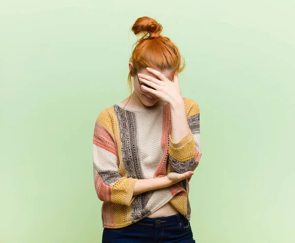 young pretty red head woman looking stressed, ashamed or upset, with a headache, covering face with hand against green wall