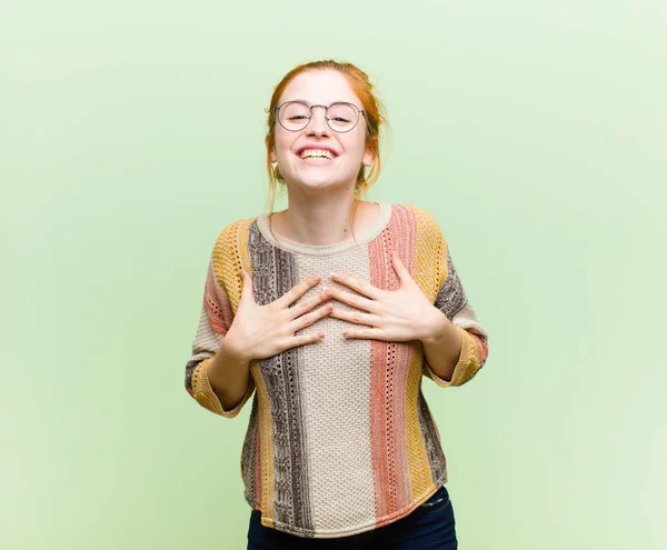 young pretty red head woman looking happy, surprised, proud and excited, pointing to self against green wall