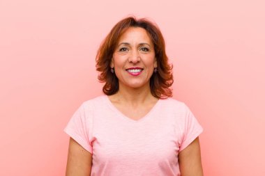Orta yaşlı kadın pozitif ve kendinden emin gülümsüyor. Tatmin olmuş, arkadaş canlısı ve pembe duvarın karşısında mutlu görünüyor.