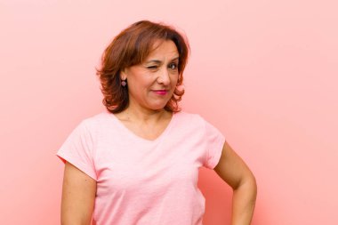 Orta yaşlı bir kadın mutlu ve arkadaş canlısı görünüyor. Pembe duvara karşı olumlu bir tavırla sana gülümsüyor ve göz kırpıyor.