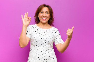 Orta yaşlı bir kadın kendini mutlu, şaşkın, tatmin olmuş ve şaşırmış hissediyor. İyi olduğunu gösteriyor ve jestler yapıyor. Mor duvara gülümsüyor.