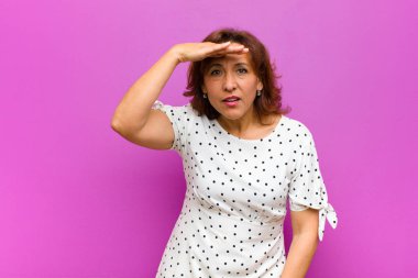 Orta yaşlı kadın şaşkın ve şaşkın görünüyor, el üstünde uzak bakıyor, izliyor ya da mor duvara karşı arıyor