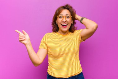 Orta yaşlı bir kadın gülüyor, mutlu, pozitif ve şaşırmış görünüyor. Mor duvara karşı yanal fotokopi alanını gösteren harika bir fikrin farkına varıyor.