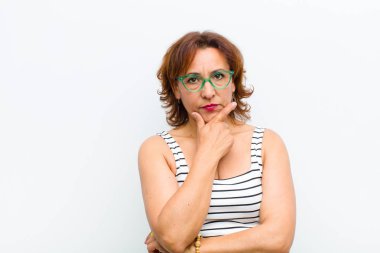 Orta yaşlı güzel bir kadın ciddi, düşünceli ve güvensiz görünüyor, bir kolu çapraz ve çene üzerinde, seçenekleri beyaz duvara karşı tartıyor