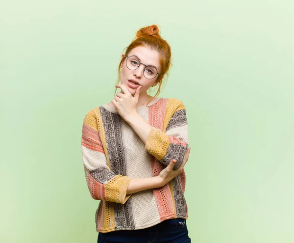 young pretty red head woman looking serious, thoughtful and distrustful, with one arm crossed and hand on chin, weighting options against green wall