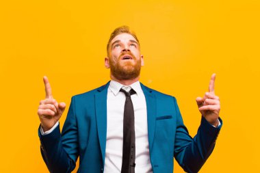 Genç kızıl saçlı işadamı şaşkın, şaşkın ve ağzı açık görünüyor, turuncu arka planda boşluğu kopyalamak için iki elini havaya kaldırıyor.