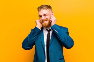 Genç kızıl saçlı işadamı sinirli, stresli ve sinirli görünüyor, her iki kulağını da turuncu arka planda sağır edici bir ses veya yüksek sesle kapatıyor.