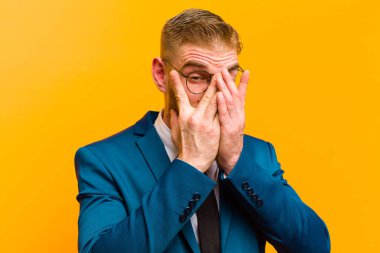 Genç kızıl saçlı işadamı korkmuş ya da utanmış hissediyor, gözleri yarı kapalı turuncu arka planda dikizliyor ya da dikizliyor.