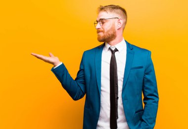 Genç kızıl saçlı işadamı mutlu ve güler yüzlü, elinde turuncu arka planda duran bir nesneye ya da konsepte bakıyor.