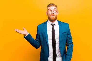 Genç kızıl saçlı işadamı şaşırmış ve şok olmuş görünüyor, çenesi düşmüş, elini turuncu arka planda açık bir nesneyi tutuyor.