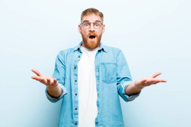 Genç kızıl saçlı adam son derece şaşkın ve şaşırmış, endişeli ve paniklemiş hissediyor, yumuşak mavi arka plana karşı stresli ve dehşetli bir bakışla