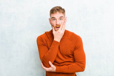 Genç, kızıl saçlı, kaplumbağa boynu takan, şok içinde ağzı açık, elleri ve kolları çaprazlanmış, beton duvara karşı sersemlemiş ve şaşırmış hisseden bir adam.