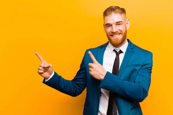 young red head businessman smiling happily and pointing to side and upwards with both hands showing object in copy space against orange background