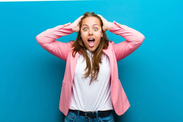 young woman raising hands to head, open-mouthed, feeling extremely lucky, surprised, excited and happy against blue background