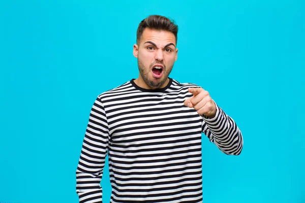 young hispanic man pointing at camera with an angry aggressive expression looking like a furious, crazy boss against blue background