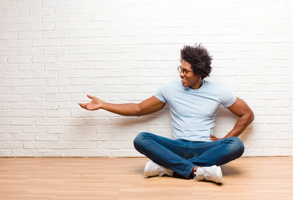 young black man feeling happy and cheerful, smiling and welcoming you, inviting you in with a friendly gesture sitting on the floor at home