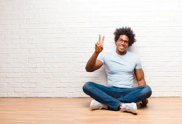 若い黒人男性は笑顔で幸せそうで気楽でポジティブで勝利や平和を手でジェスチャーで家の床に座って — ストック写真