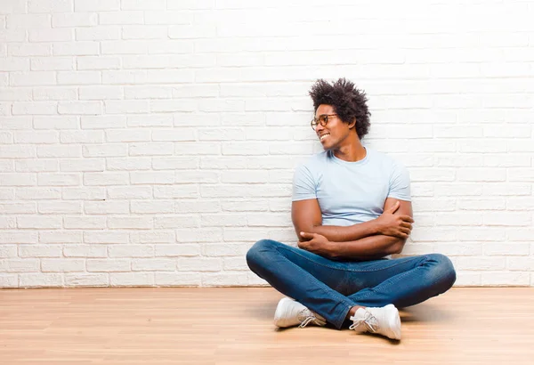 young black man smiling gleefully, feeling happy, satisfied and relaxed, with crossed arms and looking to the side sitting on the floor at home