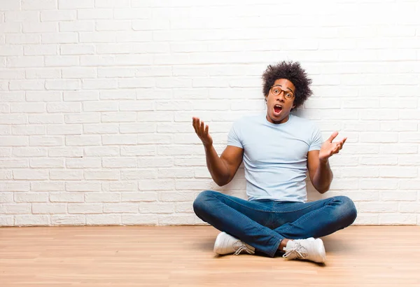 young black man open-mouthed and amazed, shocked and astonished with an unbelievable surprise sitting on the floor at home