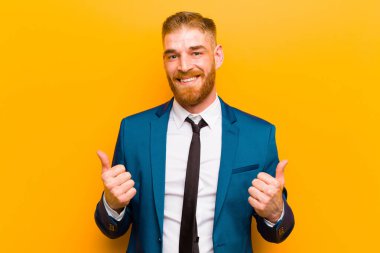 young red head businessman smiling joyfully and looking happy, feeling carefree and positive with both thumbs up against orange background clipart