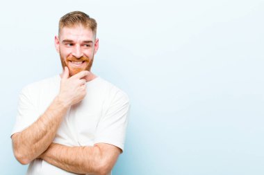 Genç kızıl saçlı adam mutlu, kendinden emin bir ifadeyle gülümsüyor, elleri çenesinde, merak ediyor ve mavi arka plana bakıyor.