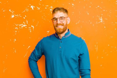 Genç kızıl saçlı adam mutlu bir şekilde gülümsüyor, eli kalçasında ve kendinden emin, pozitif, gururlu ve dost canlısı bir tavırla grunge turuncu duvarına karşı.