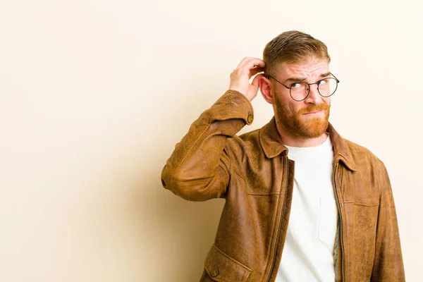 young red head man feeling puzzled and confused, scratching head and looking to the side against beige background