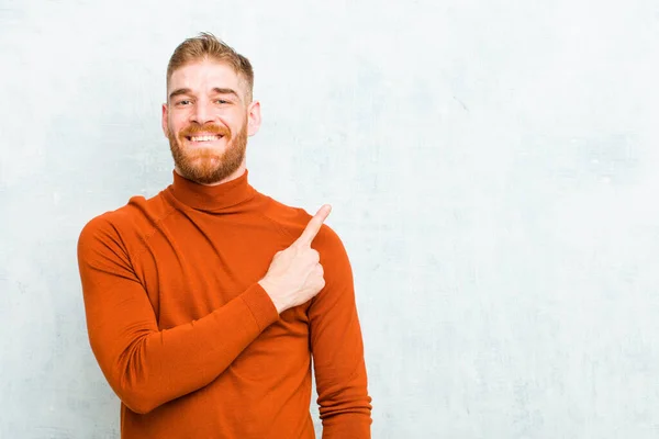 young red head man wearing turtle neck smiling cheerfully, feeling happy and pointing to the side and upwards, showing object in copy space against concrete wall