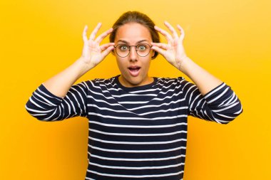 Genç, güzel bir kadın şok olmuş, şaşırmış ve şaşırmış hissediyor. Elinde turuncu arka planda hayrete düşmüş, inanmayan bir bakış var.