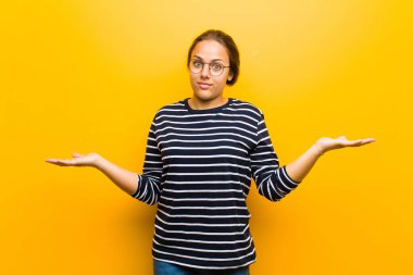 Genç güzel kadın şaşkın ve kafası karışmış hissediyor, doğru cevap ya da karardan emin değil, turuncu arka plana karşı bir seçim yapmaya çalışıyor.